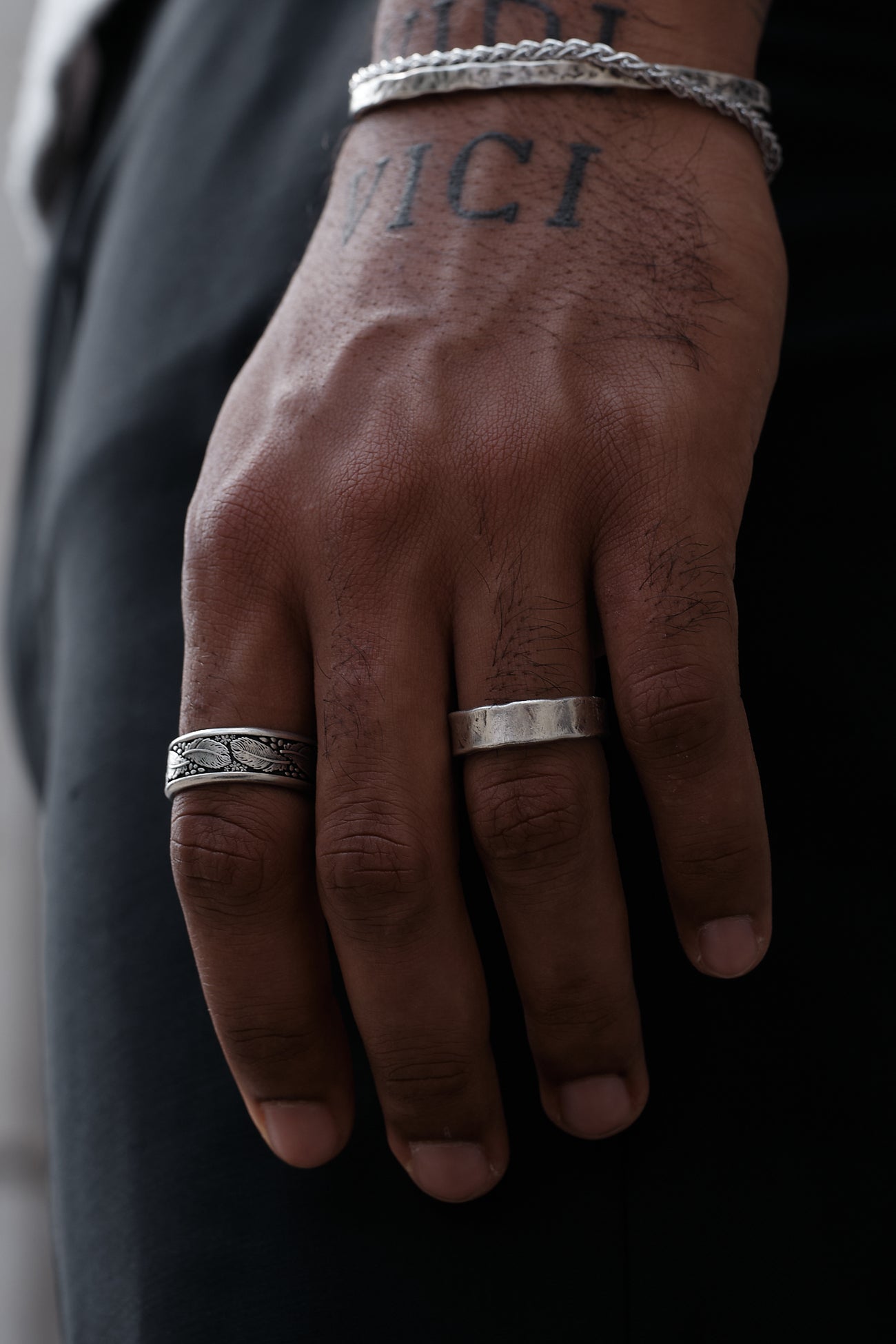 A close-up of a hand wearing silver rings and a bracelet, with a tattoo that says 'VICI.'